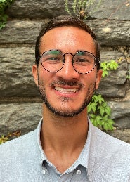 Headshot of man with glasses in a collared shirt standing in front of a stone wall.