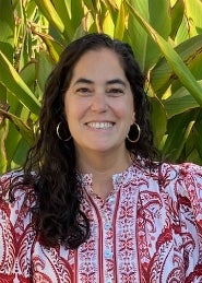 Woman with long wavy dark brown hair in red and white blouse with greenery in the background.